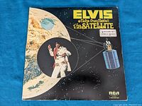 Front cover of Elvis Presley 'Aloha from Hawaii via Satellite' double vinyl album showing artwork of Elvis in white jumpsuit with microphone against space background including Earth and satellite.