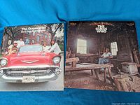 Cover of So Long Bannatyne featuring four men leaning on a red classic car outdoors, next to Share the Land album cover with a man sitting beside a wooden table in a dimly lit room.