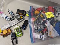 Multiple Hot Wheels cars along with a plastic airplane and car carrier trailer, some cars loose and some inside a plastic bag