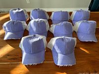 10 identical women's distressed ponytail caps arranged on a wooden surface. Caps are light purple on the front with white mesh backs and distressed visors.