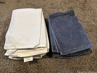 Stack of 7 white towels and 5 blue towels folded and stacked on carpeted floor.