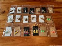 Wide shot showing all 19 sets of earrings in clear plastic packaging on card backings, arranged in rows on a wooden table.