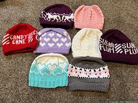 Photo showing all eight hand-knitted children's hats arranged on carpet, featuring various colors and patterns including candy cane, horses, hearts, cable knits, and text designs.