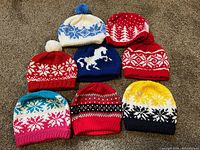 Eight assorted hand-knitted teen hats arranged on carpet showing various color patterns, pom-poms, and winter designs.