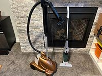 Photo showing both the Conair upright garment steamer and Bissell stick vacuum together, displayed on carpet in front of a fireplace.
