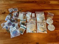 Top-down photo showing an assortment of small clear plastic bags containing various gold and silver jewelry findings such as ear hooks, jump rings, beads, and clasp components, along with multicolored metal leaves and flowers for jewelry making.