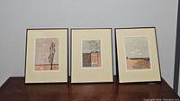 Three modern art prints displayed on a dark wood surface, showcasing full lot contents and framing