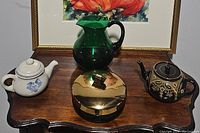 Photo showing all 4 serving pieces on wooden surface under framed artwork: white teapot, black ornate teapot, green glass pitcher, gold lidded dish