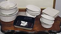 Eleven pieces of bakeware on a table, various shapes and sizes including white ceramic and one black ceramic baking dish.