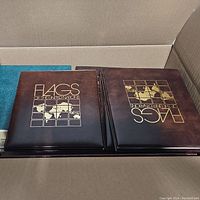 Box containing multiple stamp albums and folders, two prominent brown albums titled 'FLAGS OF THE UNITED NATIONS'