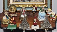Photo showing assortment of Christmas decorative pieces including metal stocking holders with Santa and Christmas trees, glass dome snow globes, ceramic bells, and Christmas village figurines.