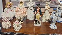 Overall assortment of nine collectible figurines displayed on a wooden surface with framed artwork background. Includes dolls, angel, soldier, and porcelain characters.