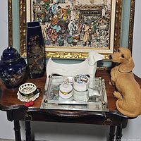 Full lot on wooden table with framed artwork background showing the white ceramic dachshund planter, a tan ceramic dog figurine, two small decorative jars on a mirrored tray, a crocheted coaster, and two blue porcelain vases with peacock designs.