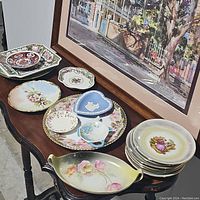 General overview of multiple decorative plates and dishes displayed on a dark wood table under framed painting