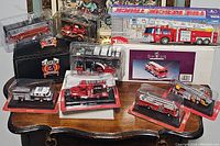 Full view of nine fire truck scale models arranged on a wooden surface, mostly in clear plastic display cases and two in retail boxes. Models are mostly red and detailed with ladders, rescue tools, and equipment.