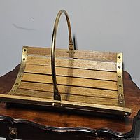 Wooden firewood basket with brass frame and arched handle placed on wooden surface.