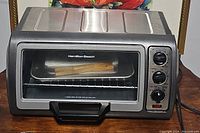 Front view of Hamilton Beach toaster oven showing stainless steel top, glass door with handle, and interior wire rack with baking tray.