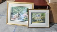 Two framed nature paintings leaning against a box on a floor, showing outdoor scenes in gold-tone frames.