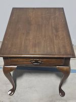 Front view of the wooden side table showing the top surface and one drawer with brass handle. Scratches and wear visible on the tabletop.