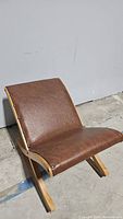 Small leather rocker chair with curved wooden base on concrete floor against gray wall, side view.