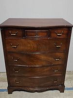 Front view of dark brown wooden five-drawer tall dresser with brass handles, showing curved drawer fronts and some surface wear.