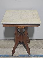 Full view of vintage wooden table with square marble top and carved detail base on wheels.
