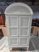 Front view of white wooden hutch with decorative fan-shaped arch and closed louvered cabinet doors.