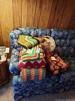 Photo of multiple folded crocheted blankets, mats, and a framed needlepoint piece stacked on a blue floral sofa