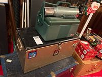 Two boxes stacked: lower wooden camping box with metal/wood details and upper Stanley plastic cooler with attached metal thermos on top.