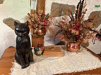 View showing painted concrete cat figurine, two copper pots with dried florals, brass bell, and wooden box with cards on cloth-covered table against textured wall.