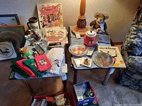 Various Disney collection items displayed on two tables including hand towels, mugs, figurines, plates, stuffed animal, LP records, a watch, necklace, and books.