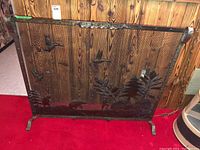 Front view of custom metal fireplace screen showing detailed nature-themed decorations including pine tree, leaves, and birds against a wooden wall background.