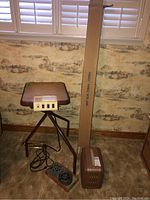 Kodak film projector on the floor connected by power cord near the Projek-Table with a textured top and control panel on front, alongside a brown portable case and a long cardboard box upright behind