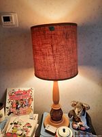 Front view of the lamp illuminated, showing the wood base and reddish textured fabric shade on a table with various surrounding items.