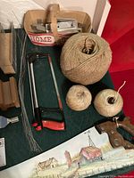 Photo showing two wooden mini miter boxes in the basket, large spool of twine, smaller twine balls, a red and black framed hacksaw, bundled wire rods, and hand painted wood saw with rustic image.