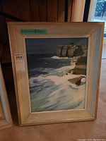Framed painting showing ocean waves and cliffs, signed by artist, with measurement label attached on frame.