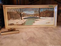 Photo of framed signed painting showing winter landscape with a frozen river and bare trees, framed in light wood with a light fitting and cord.