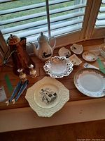 Overall view of assorted plates, teapots, and salt and pepper shakers arranged on wooden table near window with shutters.
