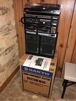 Wide angle showing entire audio system stacked vertically with original box and user manual in front, placed on carpeted floor with wood-paneled walls.