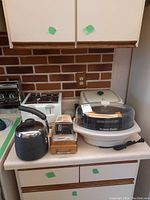 Photo showing white electric rice cooker with a transparent plastic steamer basket on top, metal teapot with black handle and wooden box-style metal grater on kitchen counter.