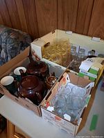 Cardboard boxes containing brown ceramic Sadler teapots and a small pot, assorted ceramic mugs, clear glass drinking glasses with gold swirl designs, and small clear glass bottles or jars.
