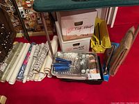 Wide view photo showing wallpaper rolls, paint trays, rollers, tile tools in boxes, new metal safety handle and rug pad in packaging, laid out on a red carpet.