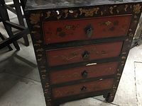 Front view of Asian style chest showing four lacquered drawers with metal drop handles and hand-painted floral designs in red and black with gold accents. Some surface wear and small chips visible.