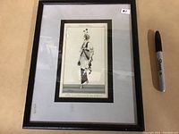 Full view of the framed "Costumes Parisiennes" print next to a black marker for scale, showing the entire piece and frame.