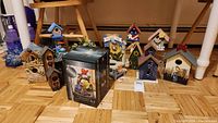 Overview of 10 decorative bird houses arranged on floor including various colorful and natural wood finishes plus one boxed item in front center.