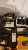 Photo shows Kodak Tourist Camera in box, Polaroid 330, TeleChallenger disc camera, Samsung film camera and other cameras arranged on table.
