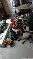 Photo shows assorted Christmas decorations on floor in basement. Visible are a wooden decoration stand, artificial Christmas tree, Santa figure, wreath, and other hanging ornaments.