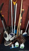 Full view of cleaning and garden tools grouped together showing mops, steamer, shovels, broom, cutter, and two small bottles of liquids on floor.