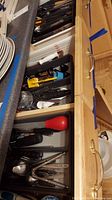 Top view of three wooden drawers open showing assorted cutlery and kitchen utensils in plastic trays, including various spoons, forks, knives, a red handled utensil, and several kitchen tools.