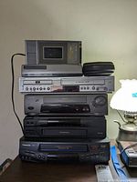 Front view of stacked VHS and VHS DVD players including rewinder units on top and beside the stack, showing brand names and model numbers.